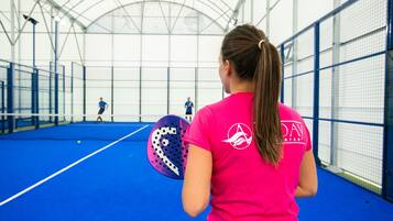 An indoor sports facility with a blue floor, a netted wall, and a person in a pink shirt holding a paddle.
