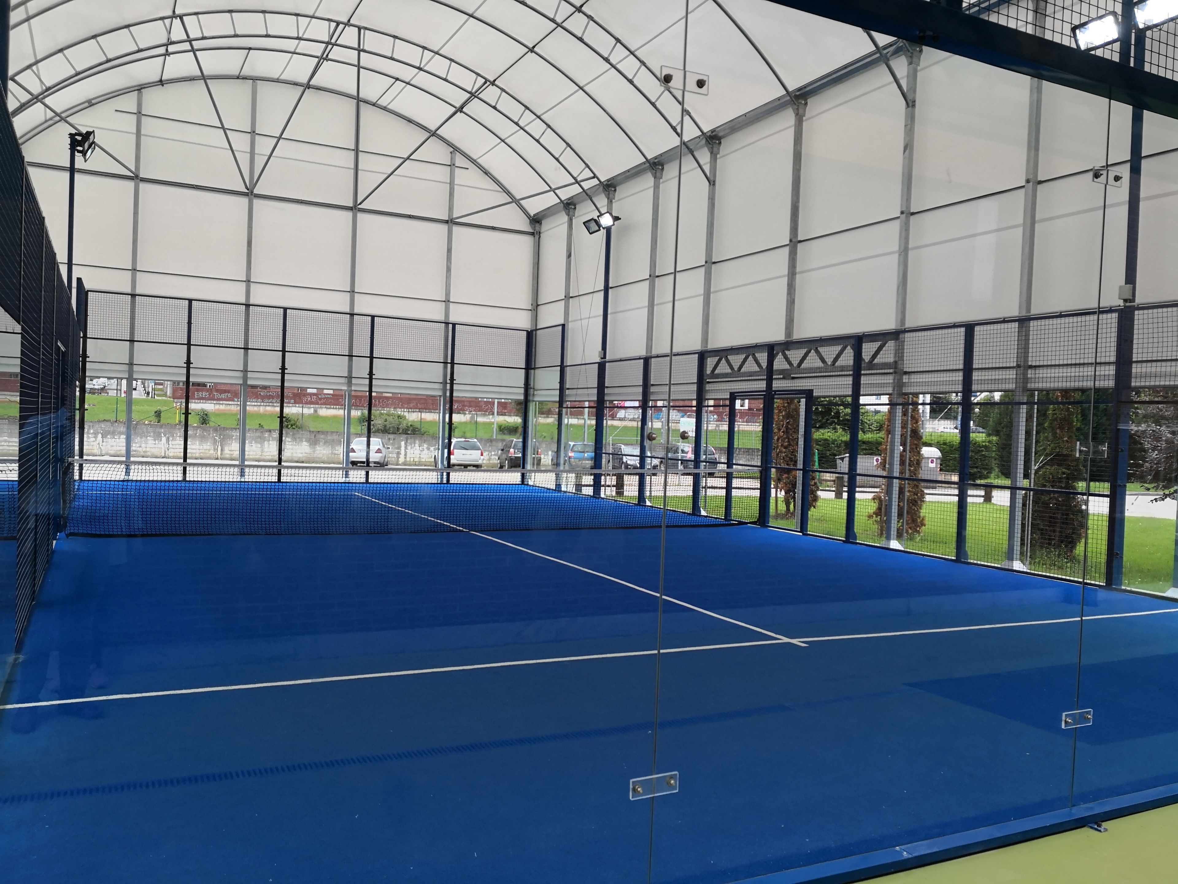 Un court de tennis intérieur, doté d’un filet, entouré d’une clôture métallique et de panneaux de verre.