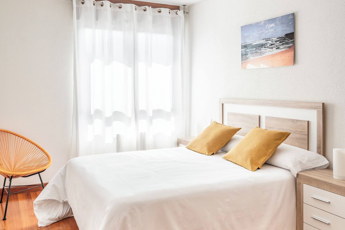 A neatly arranged bedroom with a bed, a chair, a bedside table, and a painting on the wall.
