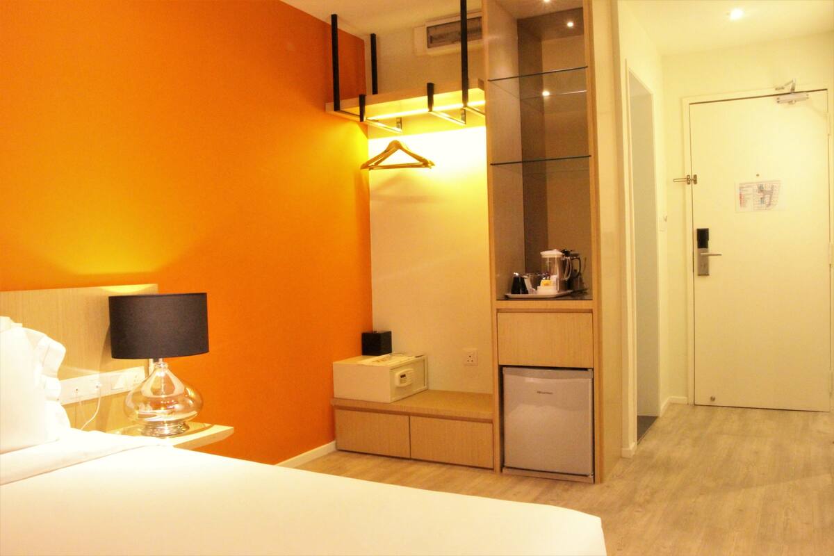 A hotel room with a bed, bedside table, lamp, and a small kitchenette area with a refrigerator.