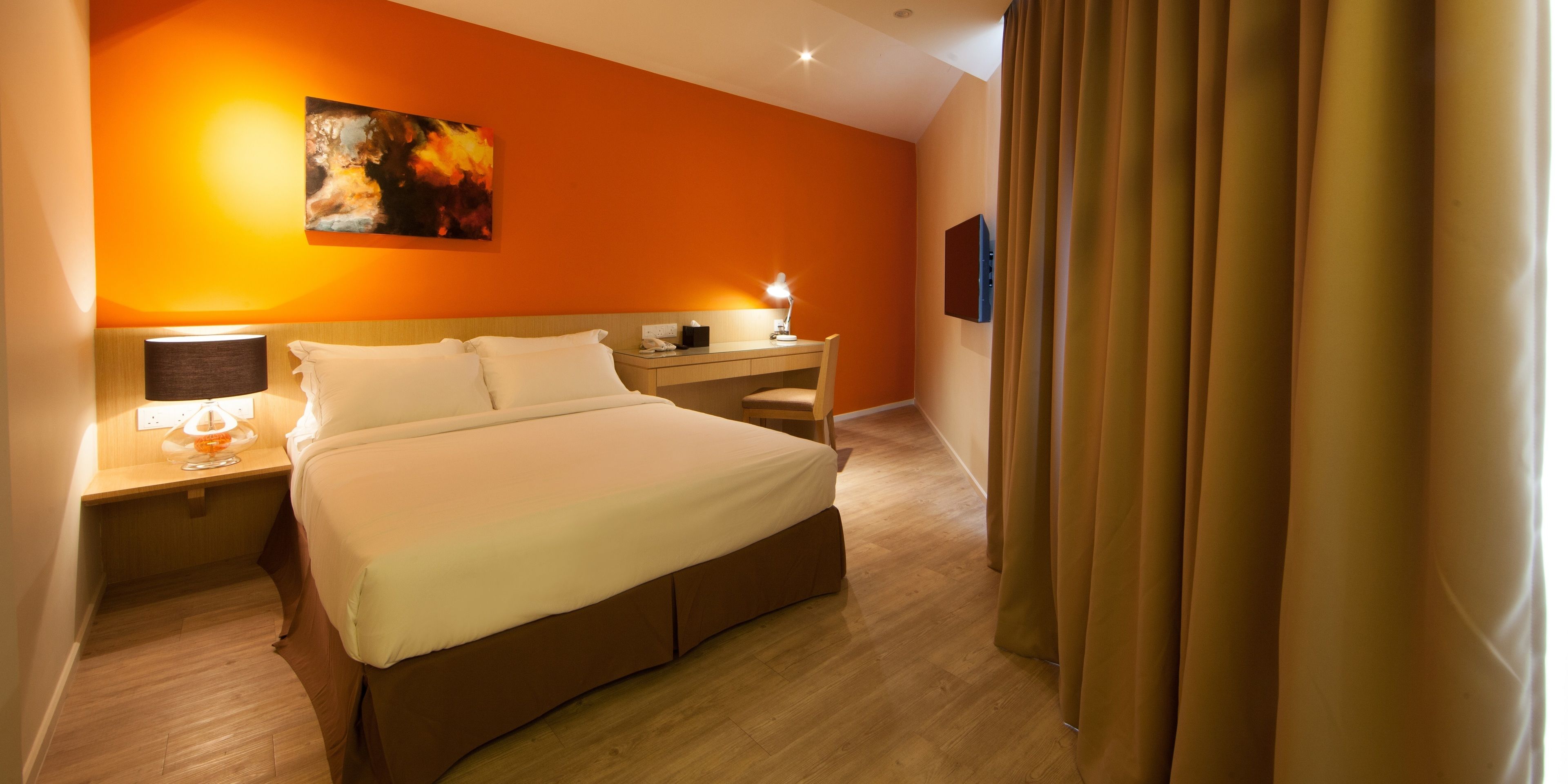 A hotel room with a large bed, a bedside table with a lamp, a desk, and a painting on the wall.