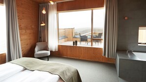 A modern hotel room with a large window, wooden walls, a bed, a chair, and a bathroom with a sink and mirror. - Fosshotel Myvatn (Myvatn)