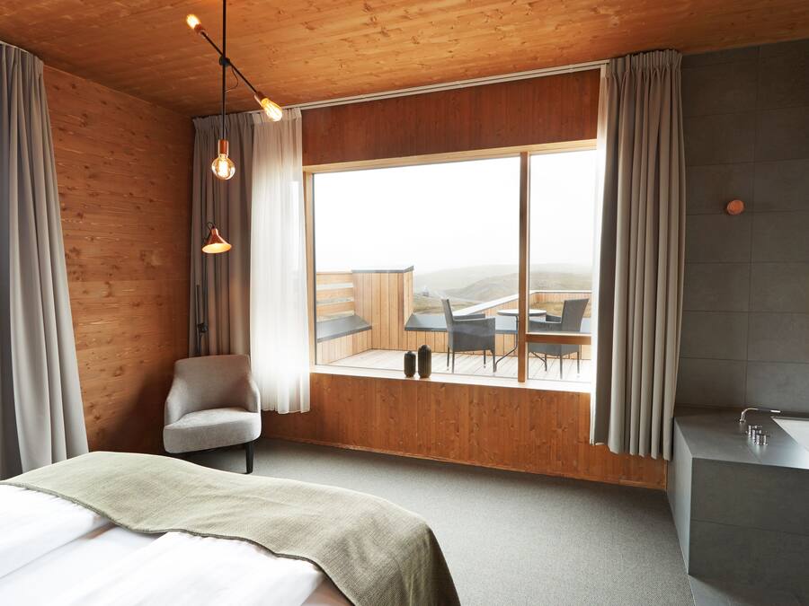 A modern hotel room with a large window, wooden walls, a bed, a chair, and a bathroom with a sink and mirror.
