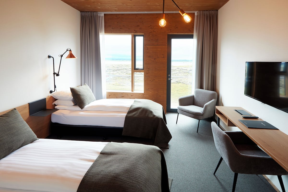 A hotel room with two beds, a desk, a chair, a TV, and a large window with a view.