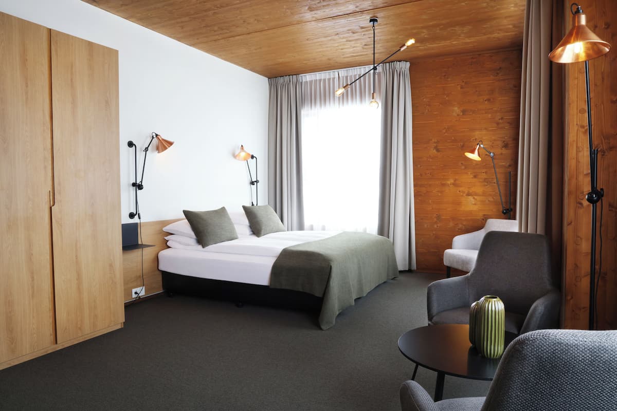 A modern hotel room with a wooden ceiling, a large bed with a green blanket, two bedside lamps, a grey armchair, a small table with a vase, and a window with curtains.