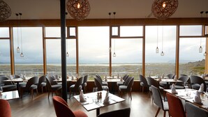 A restaurant with tables set for dining, large windows offering a view of the landscape, and hanging light fixtures. - Fosshotel Myvatn (Myvatn)
