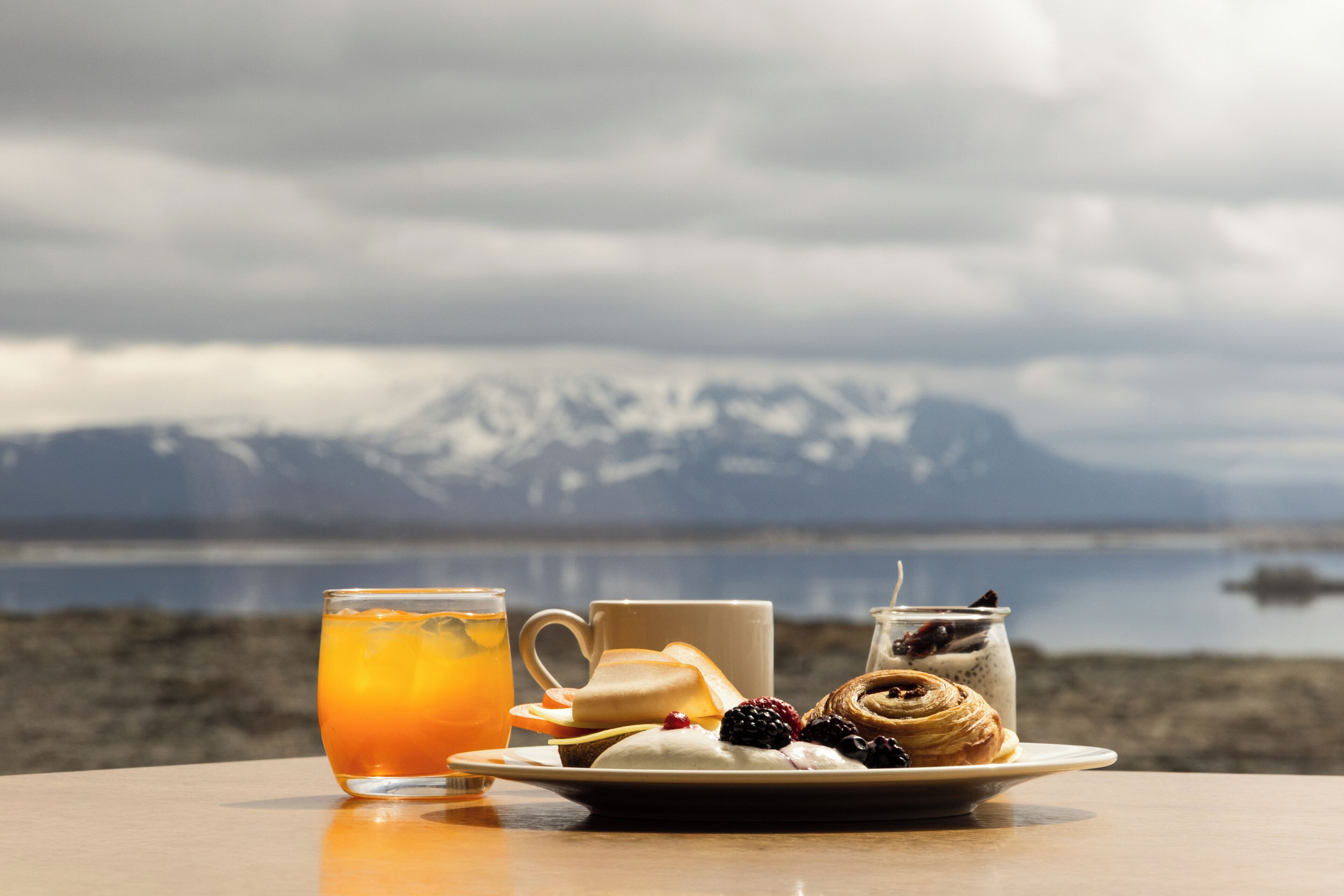 Una mesa con un plato de comida, un vaso de jugo de naranja y una taza, frente a una vista panorámica de montañas y un cuerpo de agua.