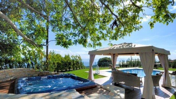 Een hottub met een stenen rand, een pergola met witte gordijnen en een zwembad op de achtergrond.