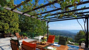 Een eethoek buiten met een pergola, houten stoelen, een gedekte tafel met borden en een fruitschaal, en uitzicht op het landschap.