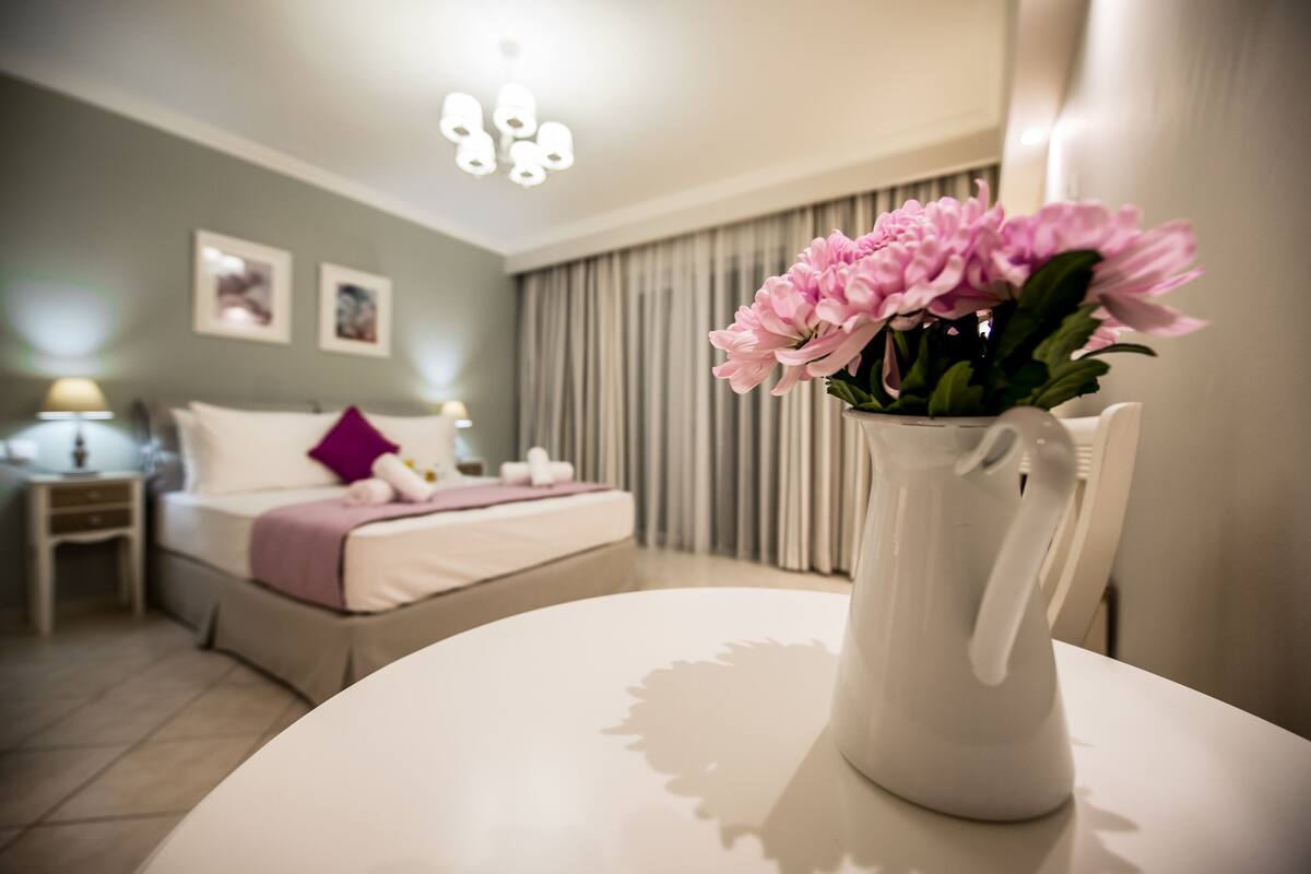 Una habitación de hotel con una cama, una mesita de noche, una mesa blanca con un jarrón de flores y una araña.