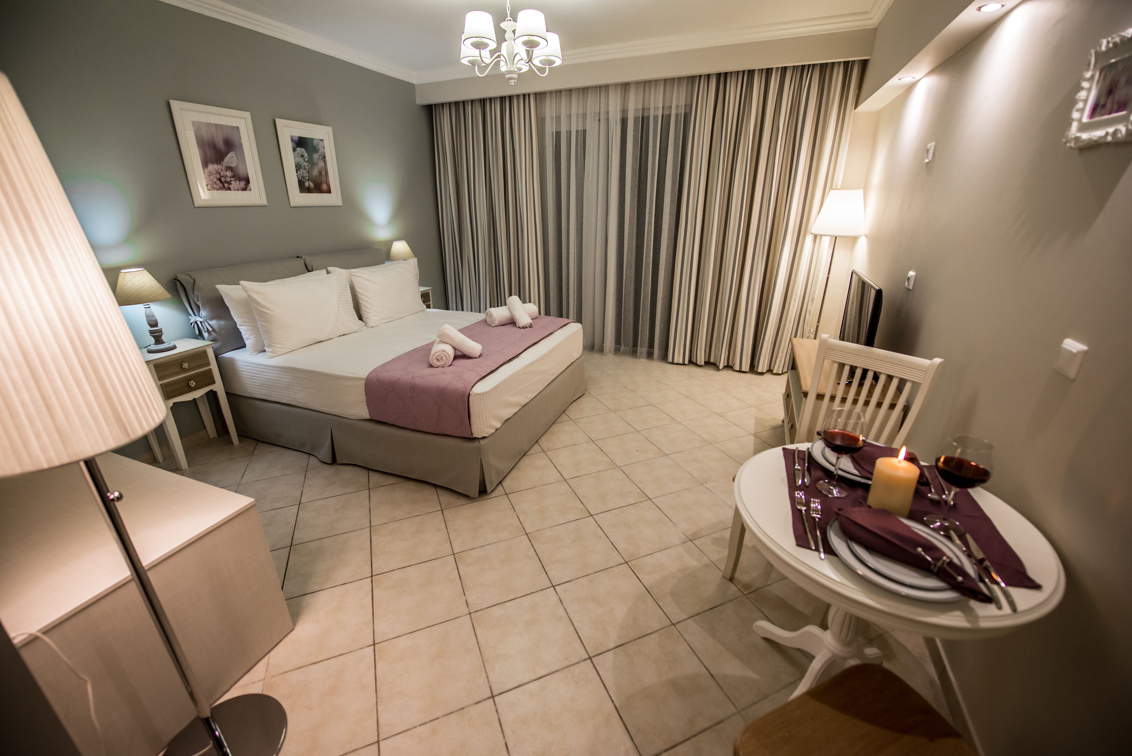 A hotel room with a bed, a bedside table with a lamp, a chair, and a small table set for dining with a candle and wine glasses.