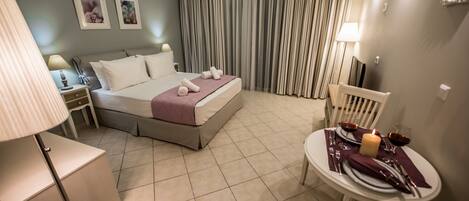 A hotel room with a bed, a bedside table with a lamp, a chair, and a small table set for dining with a candle and wine glasses.