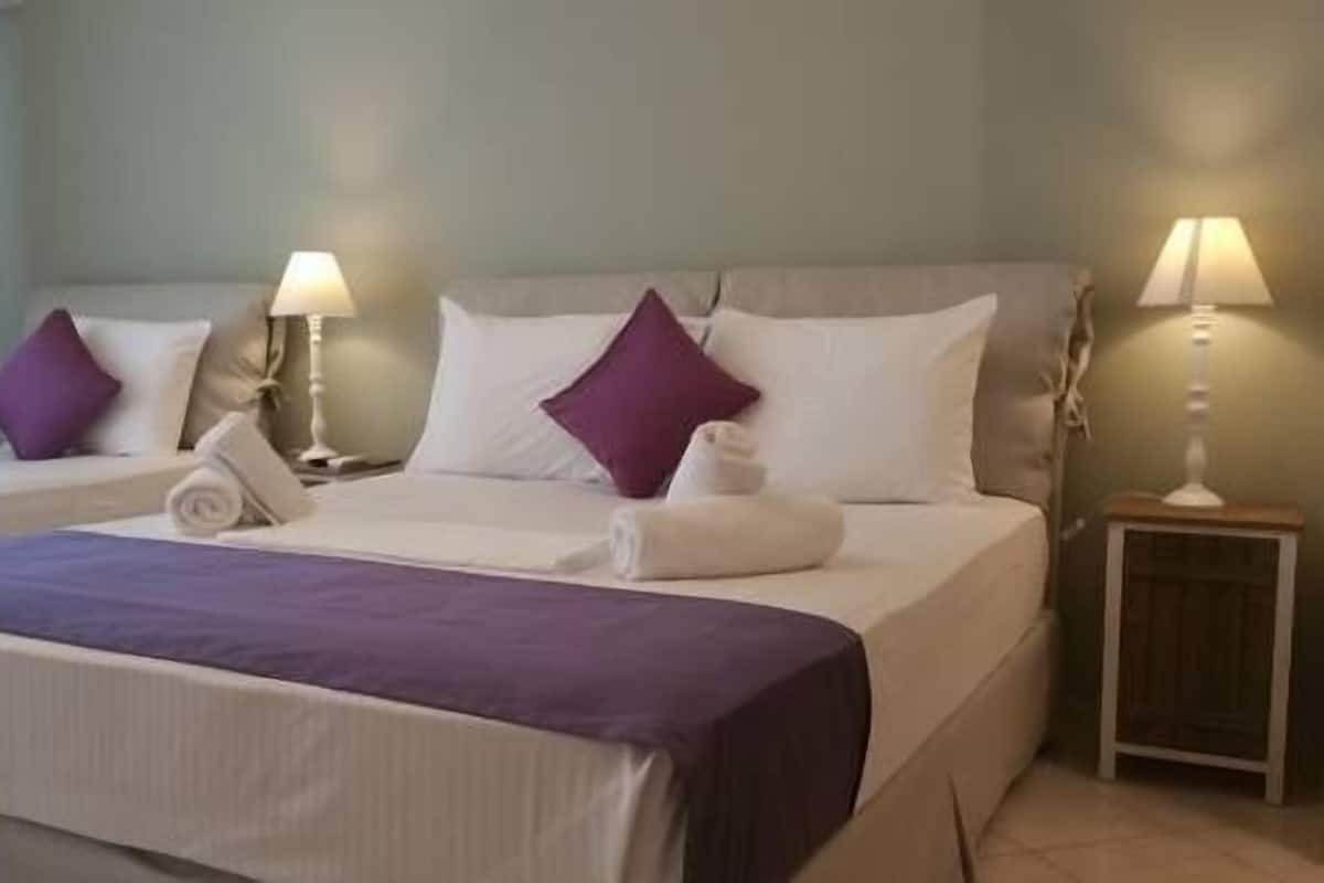 Habitación de hotel con dos camas, cada una con ropa de cama blanca y detalles morados. Hay dos lámparas de noche y una mesita de noche de madera con una lámpara.