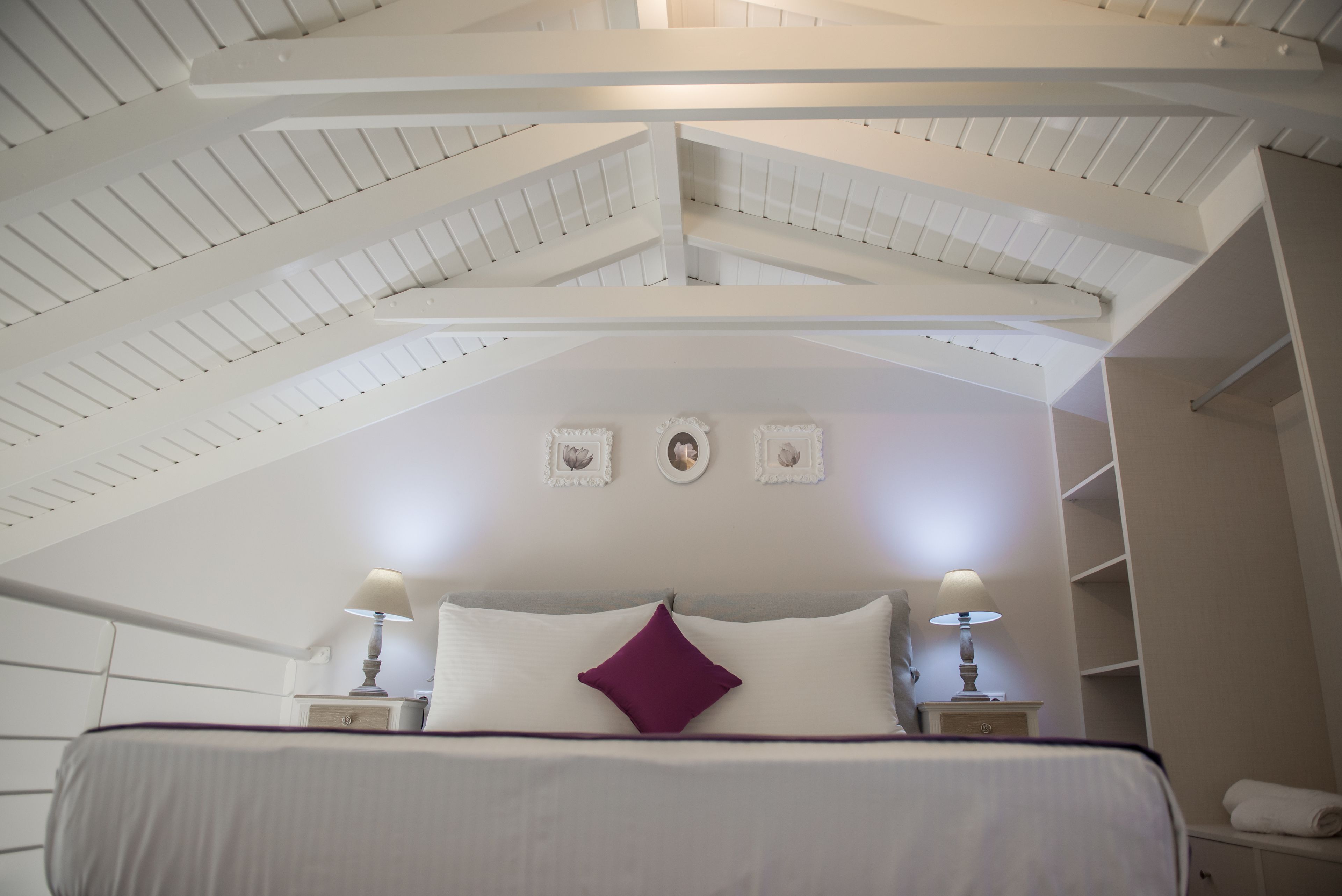 Una camera da letto con soffitto spiovente, un letto con biancheria bianca e viola, due abat-jour e un comodino.