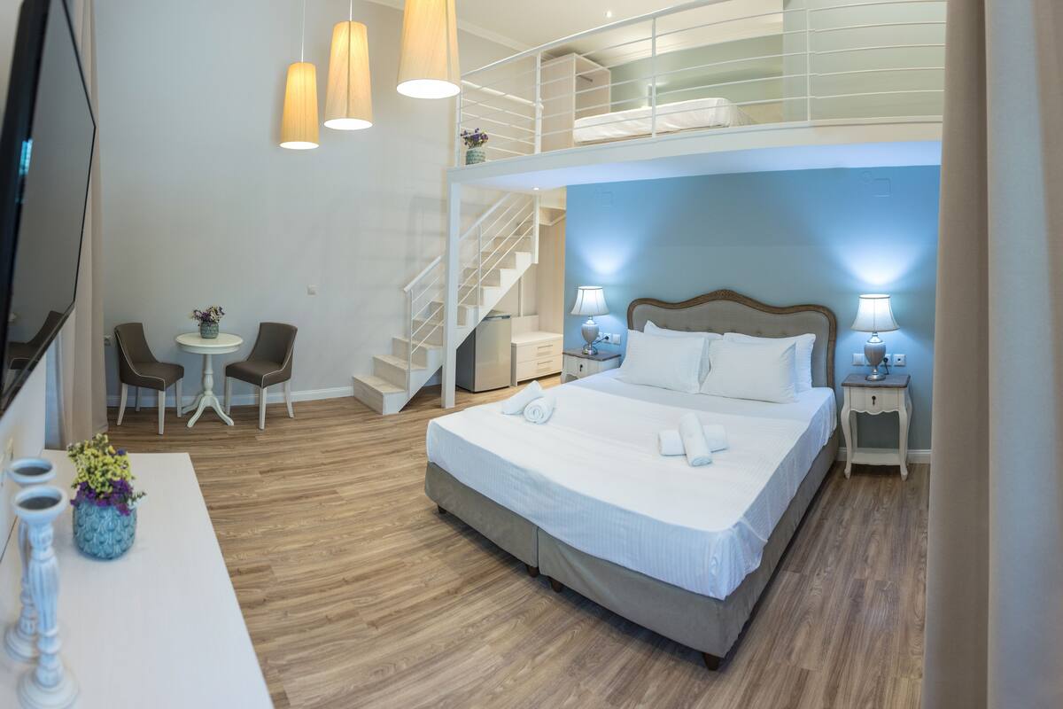 Habitación de hotel con una cama grande, un escritorio con una silla, una pequeña mesa con sillas, una escalera y un televisor.
