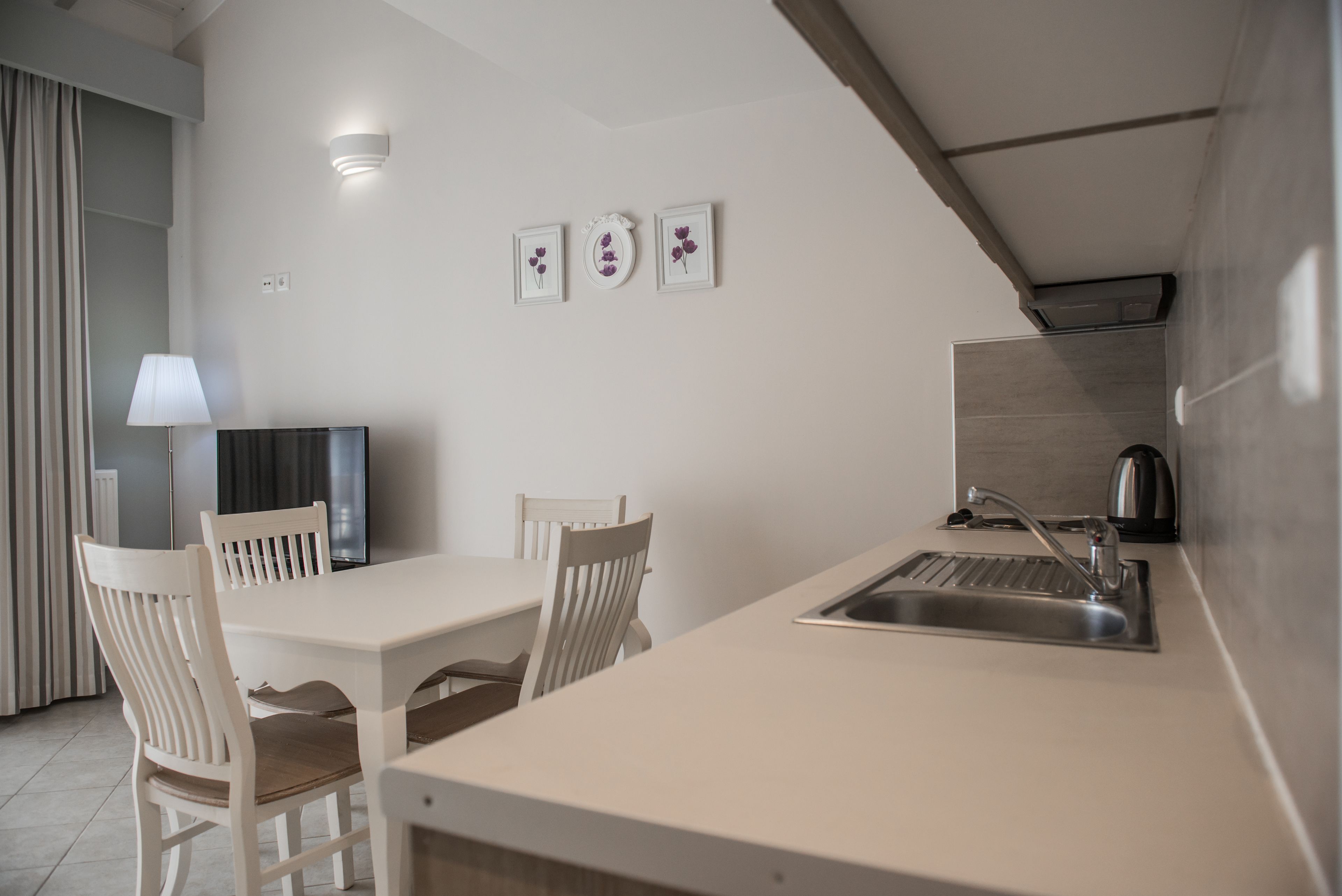 A modern kitchen with a white countertop, a sink, and a dining area with white chairs. There are framed pictures on the wall and a lamp on the left.