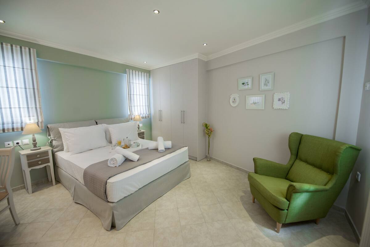 Un dormitorio ordenado con cama, mesitas de noche, un sillón verde y cuadros en la pared.