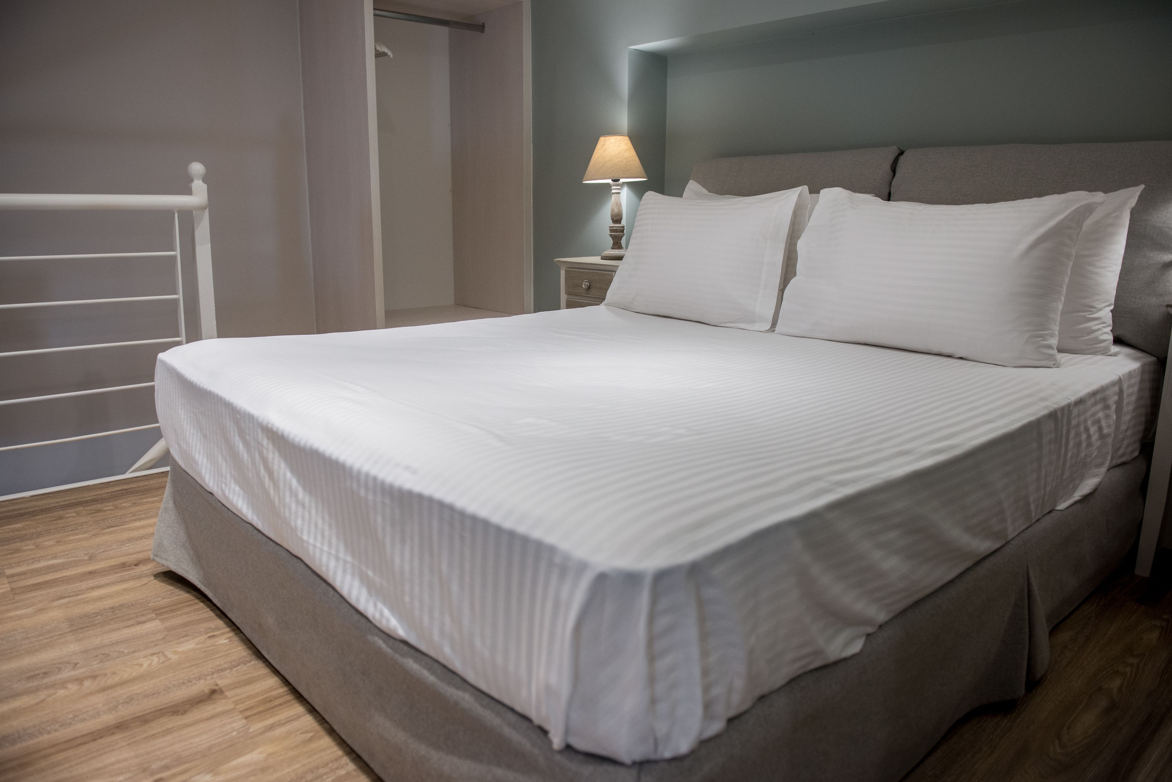 Uma cama bem arrumada com colcha e almofadas brancas, uma mesa de cabeceira com abajur e piso de madeira.