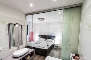 Une chambre moderne dotée d’une porte coulissante à vitres, d’un grand lit avec du linge de lit blanc, d’une table de chevet et d’une coiffeuse intégrée avec un miroir et un lavabo.