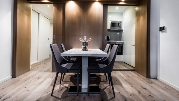 Uma sala de jantar moderna com mesa e cadeiras, um painel de parede em madeira e uma cozinha visível através de uma porta adjacente.