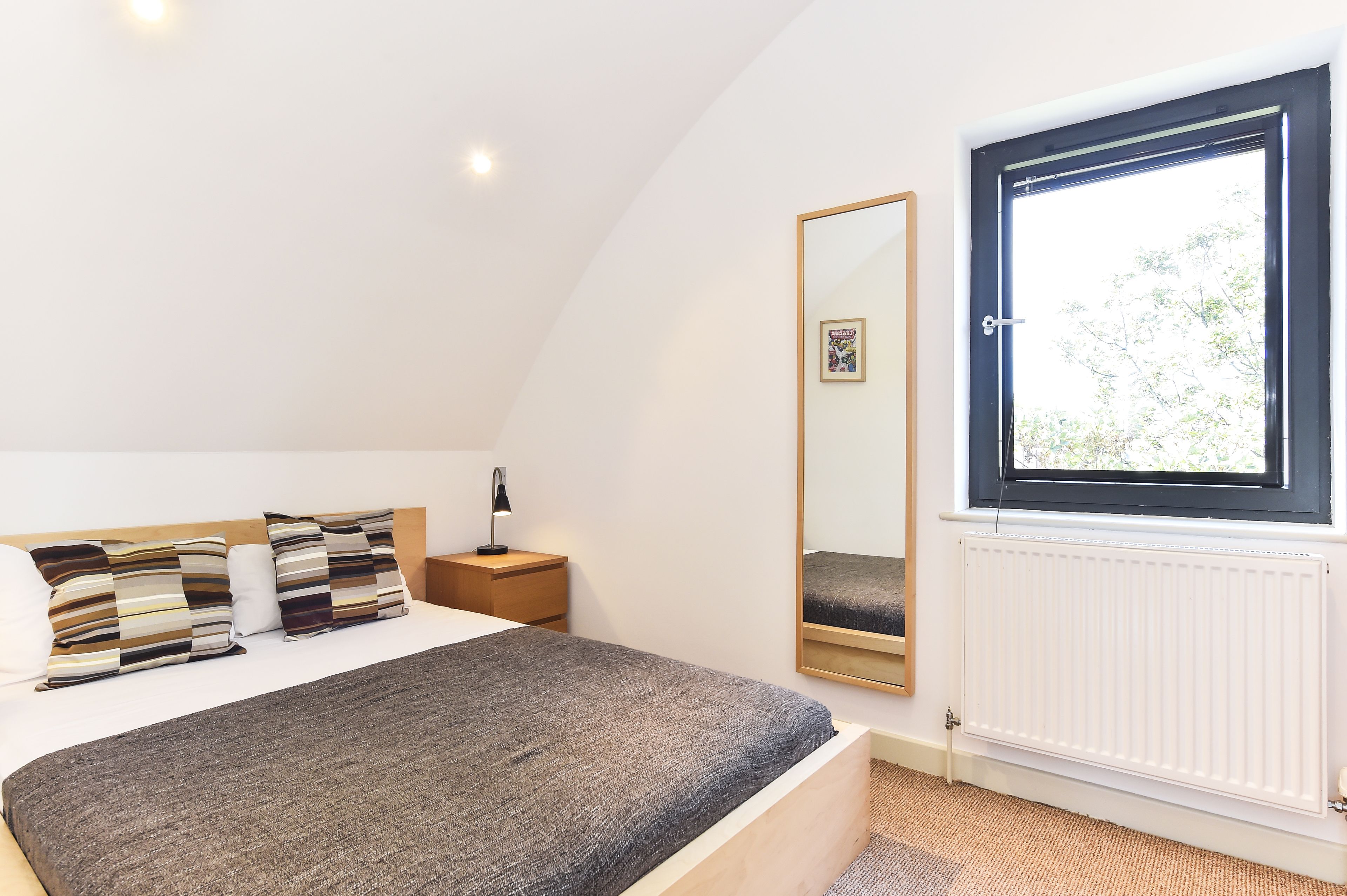 A bedroom with a large bed, a bedside table with a lamp, a window with a view of trees, and a radiator.