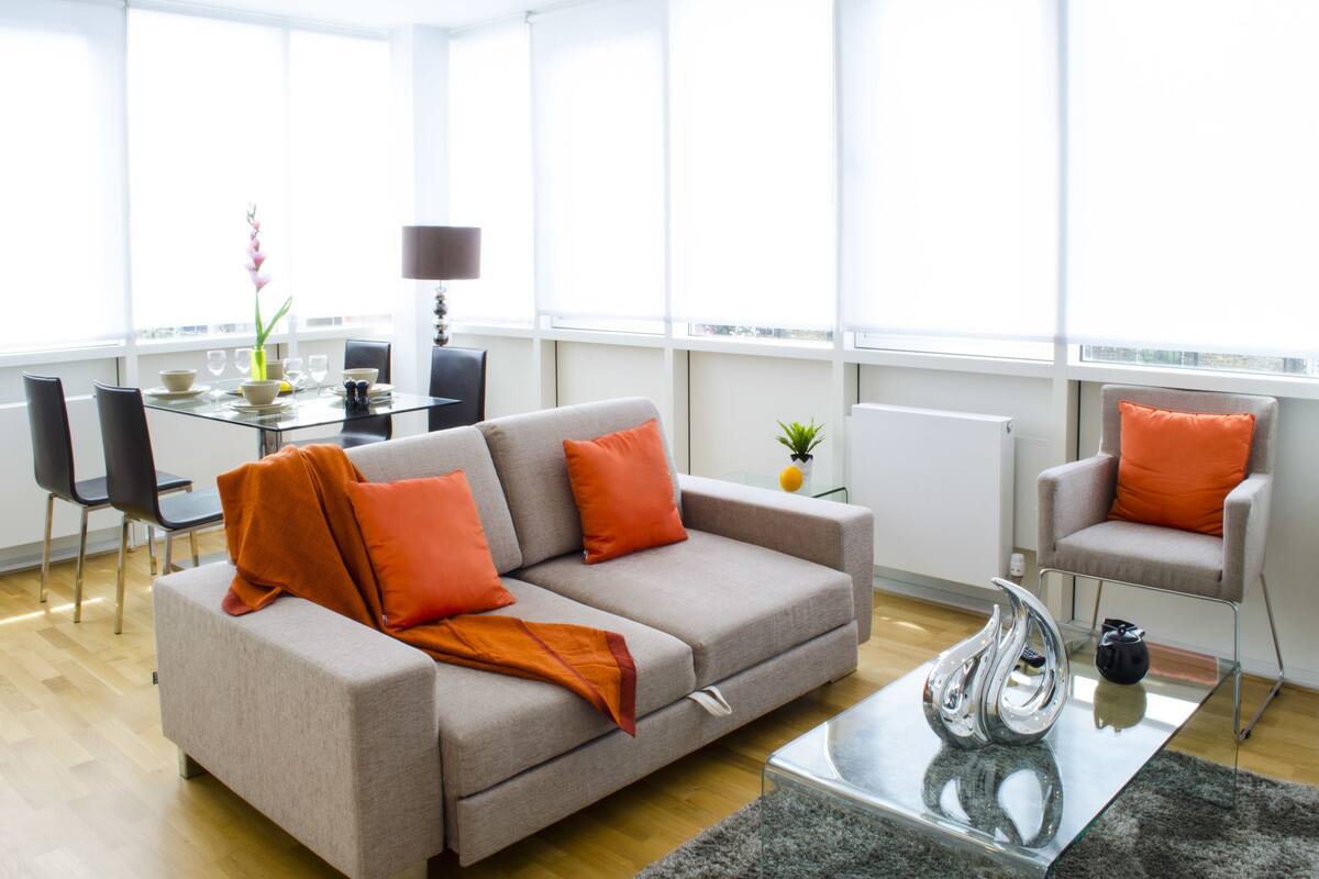 A modern living room with a grey sofa, orange cushions, a glass coffee table, and a dining area with a glass table and chairs.