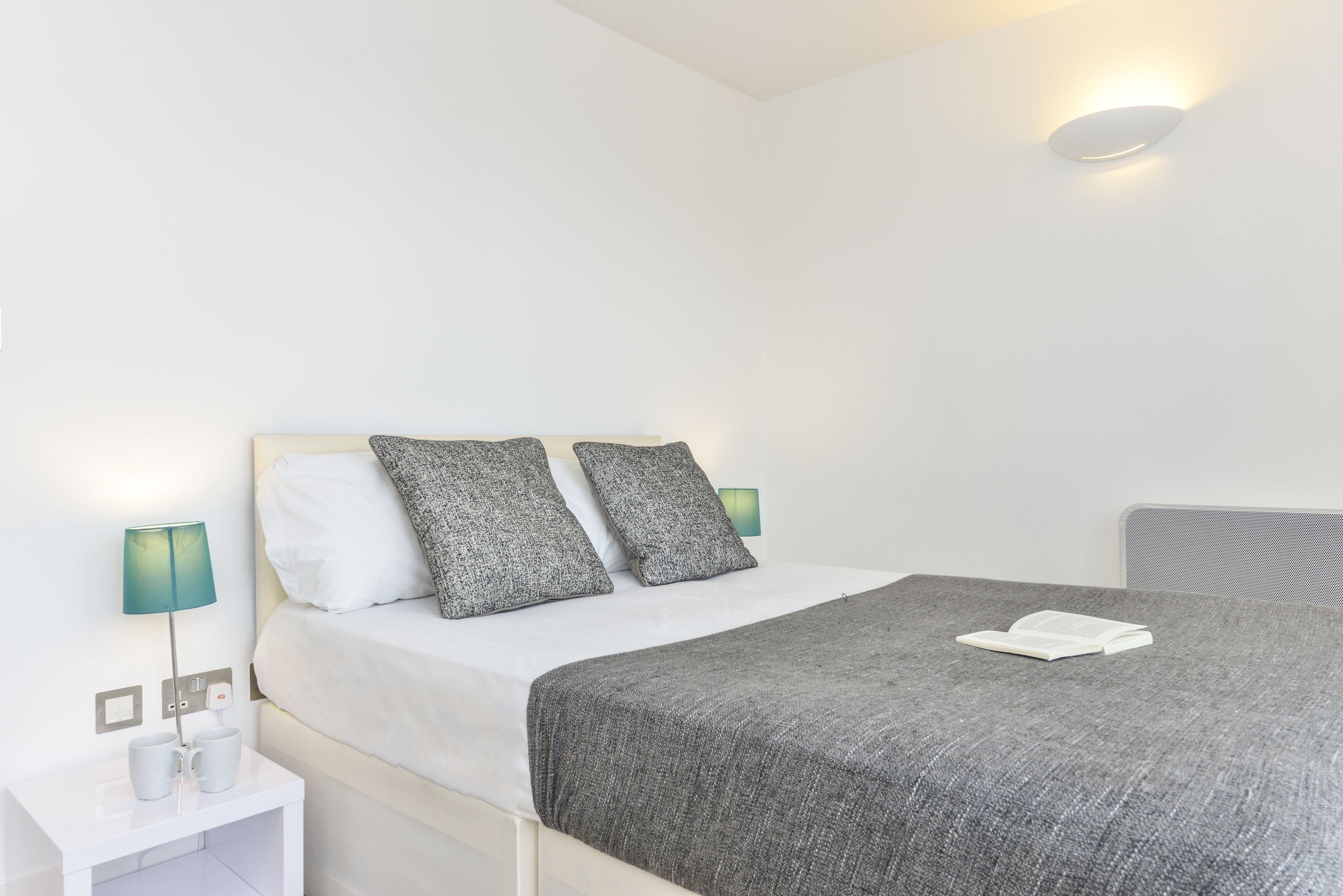 A neatly made bed with a grey blanket and two pillows, a bedside table with a lamp, and a wall-mounted light fixture.