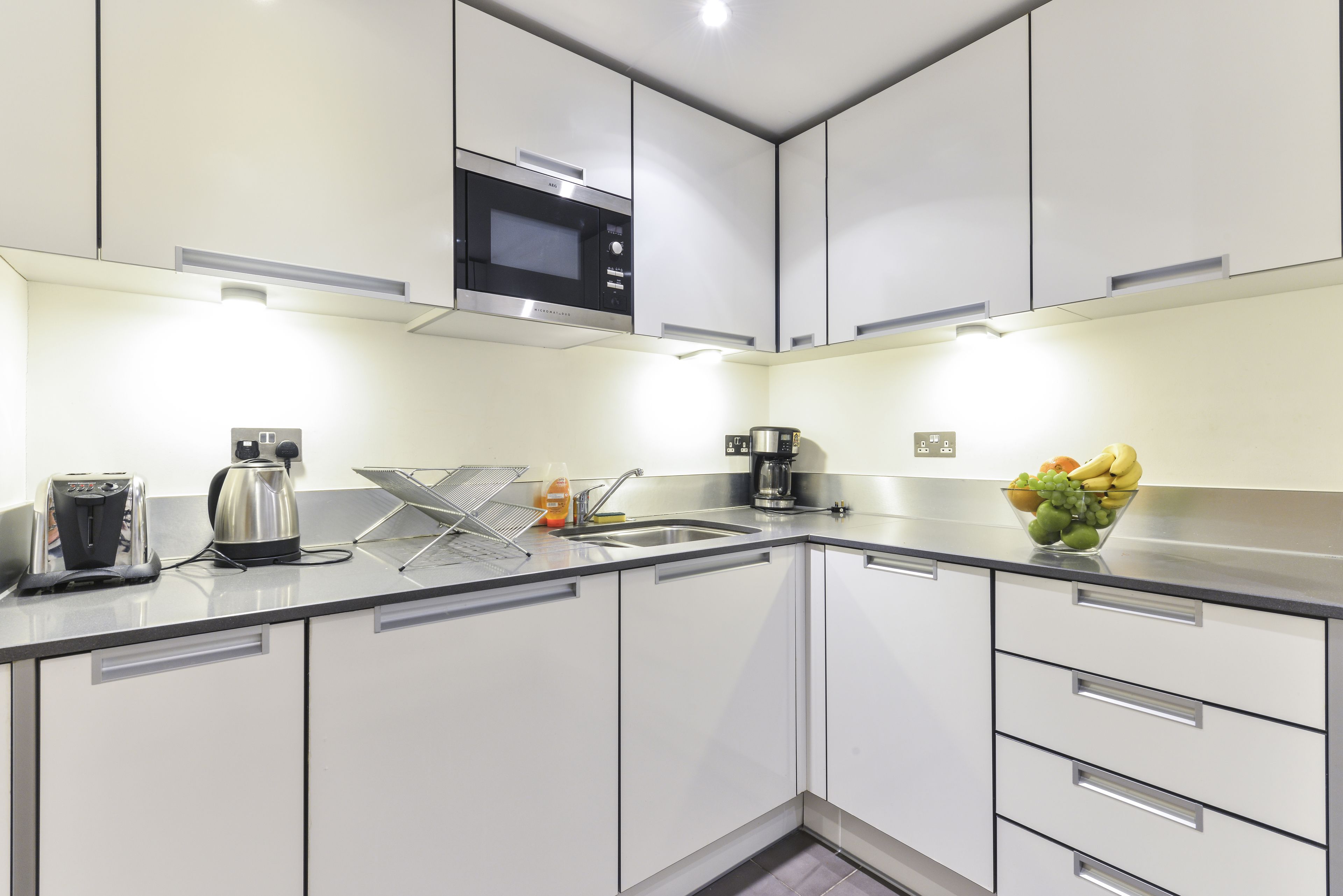 A modern kitchen with white cabinets, a stainless steel sink, a microwave, and a fruit bowl on the counter.