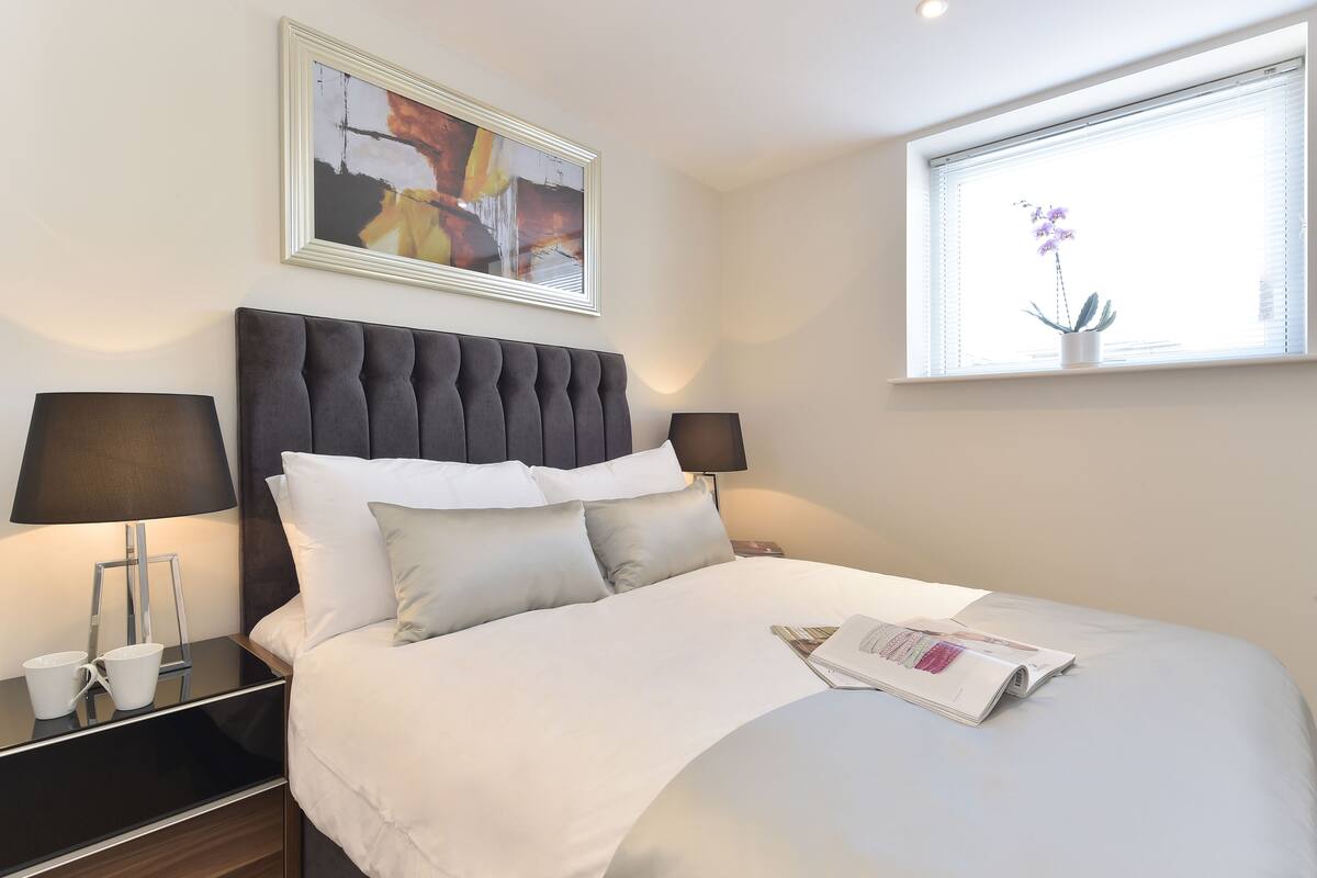 A modern hotel room with a large bed, two bedside lamps, a framed abstract painting, and a window with a potted orchid.