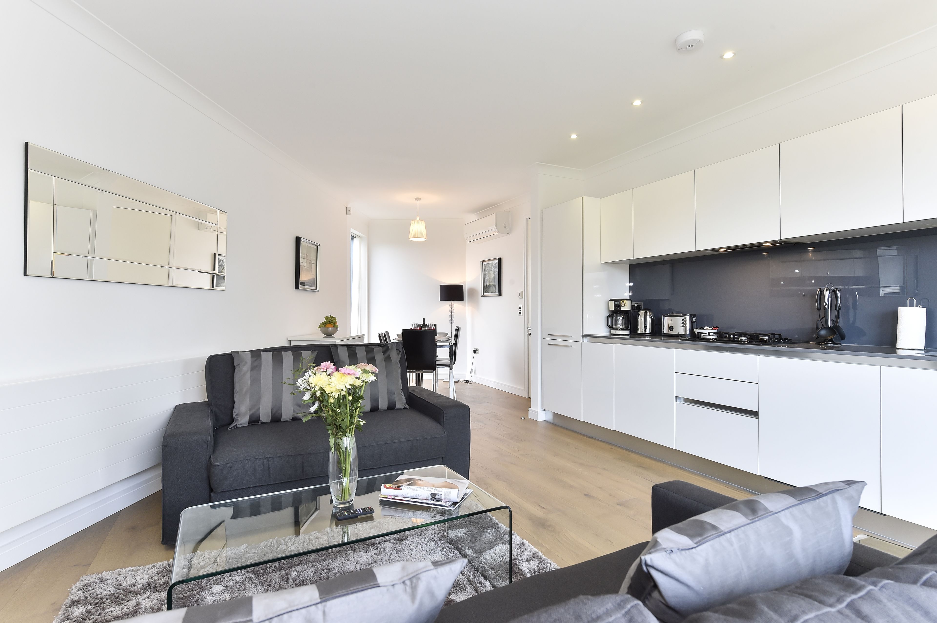 A modern apartment with a living room, a glass coffee table, a sofa, a kitchen with white cabinets, and a dining area.
