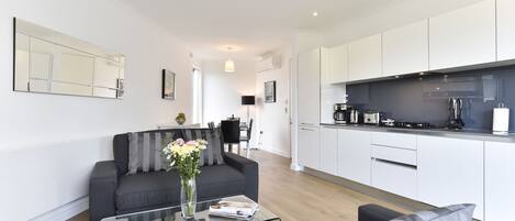 A modern apartment with a living room, a glass coffee table, a sofa, a kitchen with white cabinets, and a dining area.