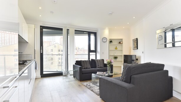 A modern living room with a sofa, a coffee table, and a TV. There is a balcony with a railing and a city view. - Tower Bridge City Apartments (London)