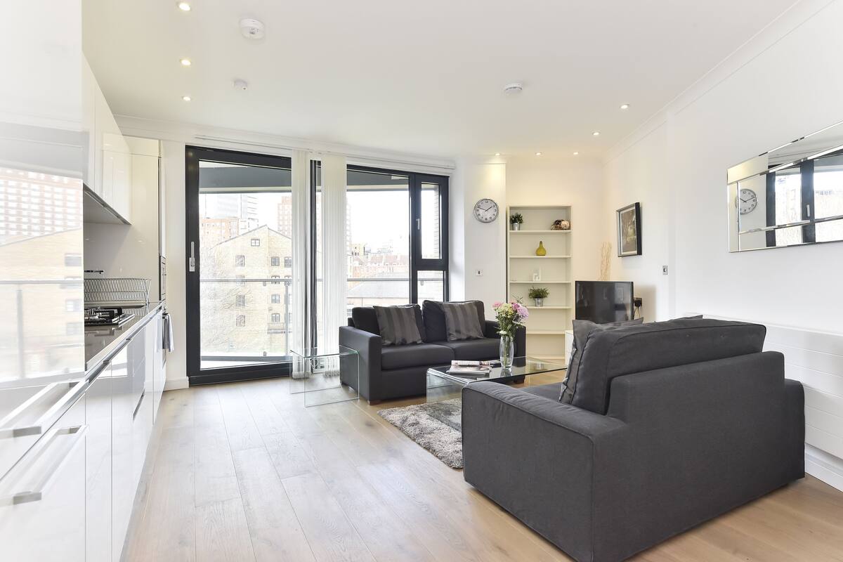 A modern living room with a sofa, a coffee table, and a TV. There is a balcony with a railing and a city view.