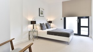 A modern bedroom with a white bed, two bedside tables with lamps, a framed picture on the wall, and a large window with a view. - Tower Bridge City Apartments (London)