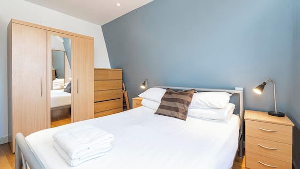 Ein ordentlich bezogenes Bett mit weißen Leinen und einem braun gestreiften Kopfkissen, ein Holzschrank und ein Nachttisch mit einer Lampe.
