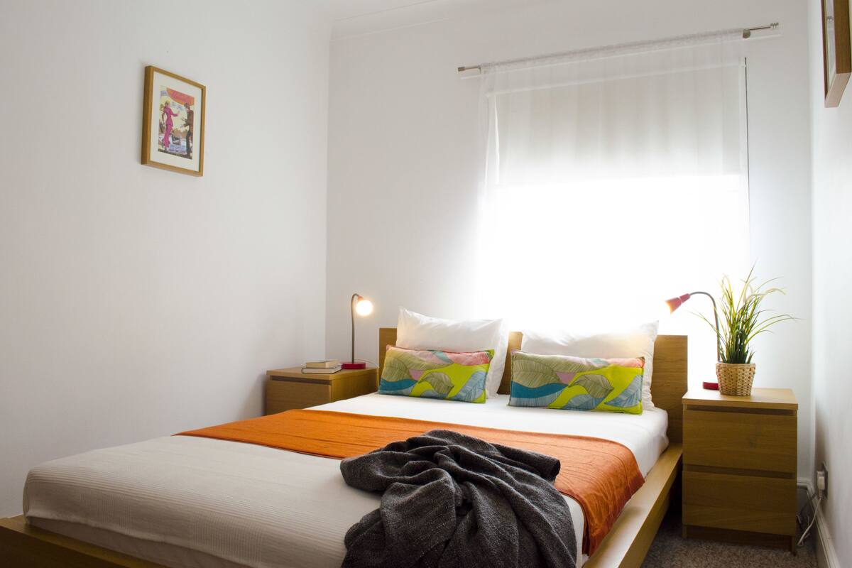 Una cama bien hecha con un edredón colorido, una lámpara de noche y una mesita con una planta.