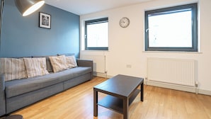 Ein modernes Wohnzimmer mit einem grauen Sofa, einem Holztisch und einer Stehlampe. Zwei Fenster mit Jalousien, eine Wanduhr und eine Heizung sind ebenfalls vorhanden.