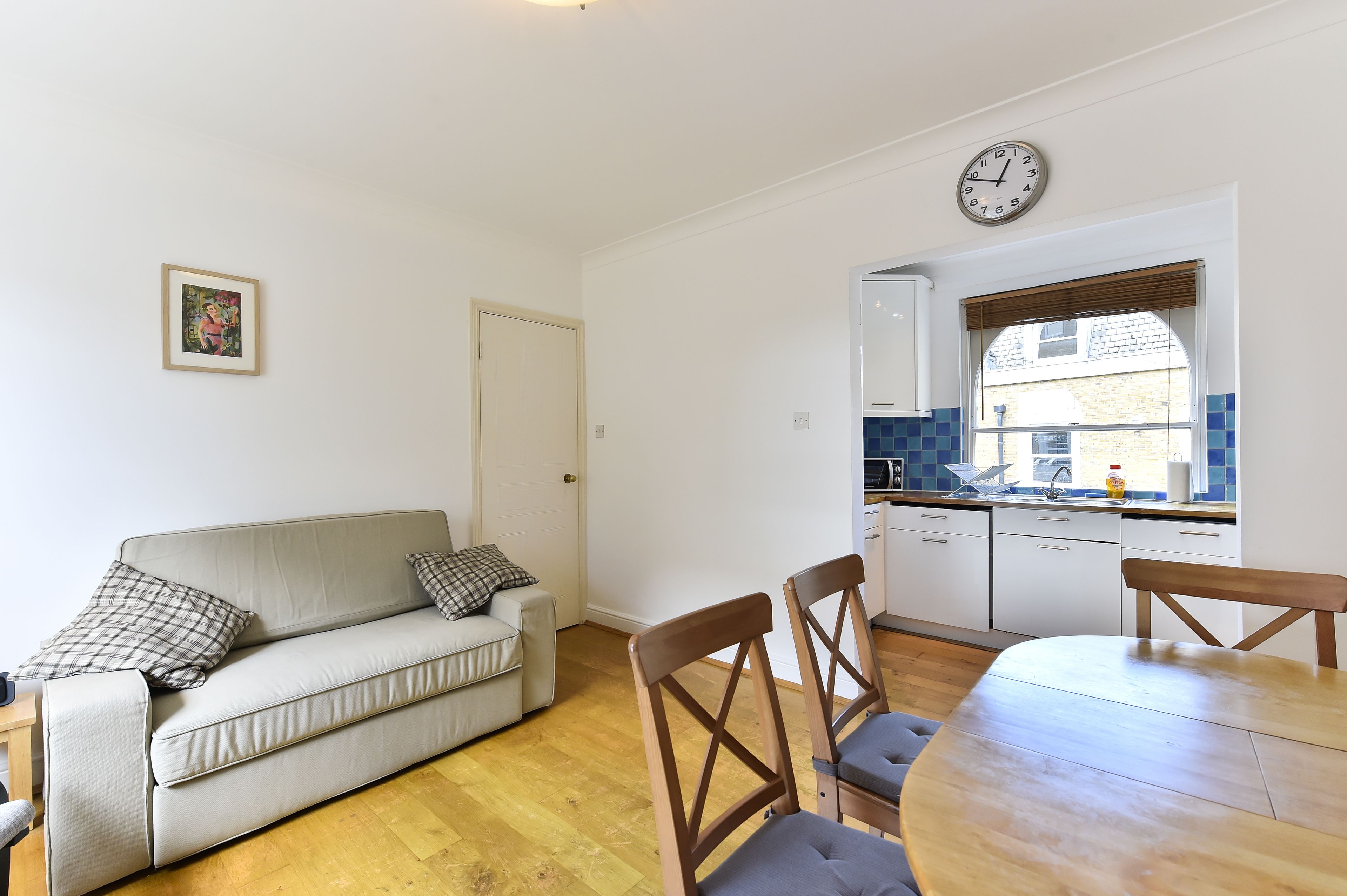 A living room with a sofa, two chairs, a dining table, a kitchen with a window, and a wall clock.