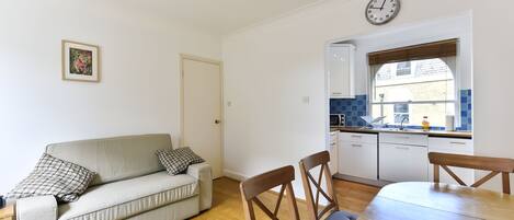 A living room with a sofa, two chairs, a dining table, a kitchen with a window, and a wall clock.