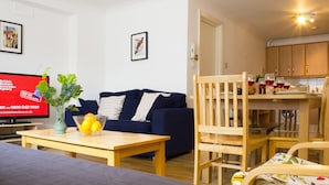 Ein Wohnzimmer mit blauem Sofa, einem Holzes Esstisch und einer Küchenzeile mit Holzschränken und einem Esstisch.