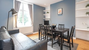 Ein modernes Wohnzimmer mit einem dunkelbraunen Ledersofa, einem Esstisch mit Stühlen, einem Fernseher und einbauartigem Bücherregal mit Pflanzen.