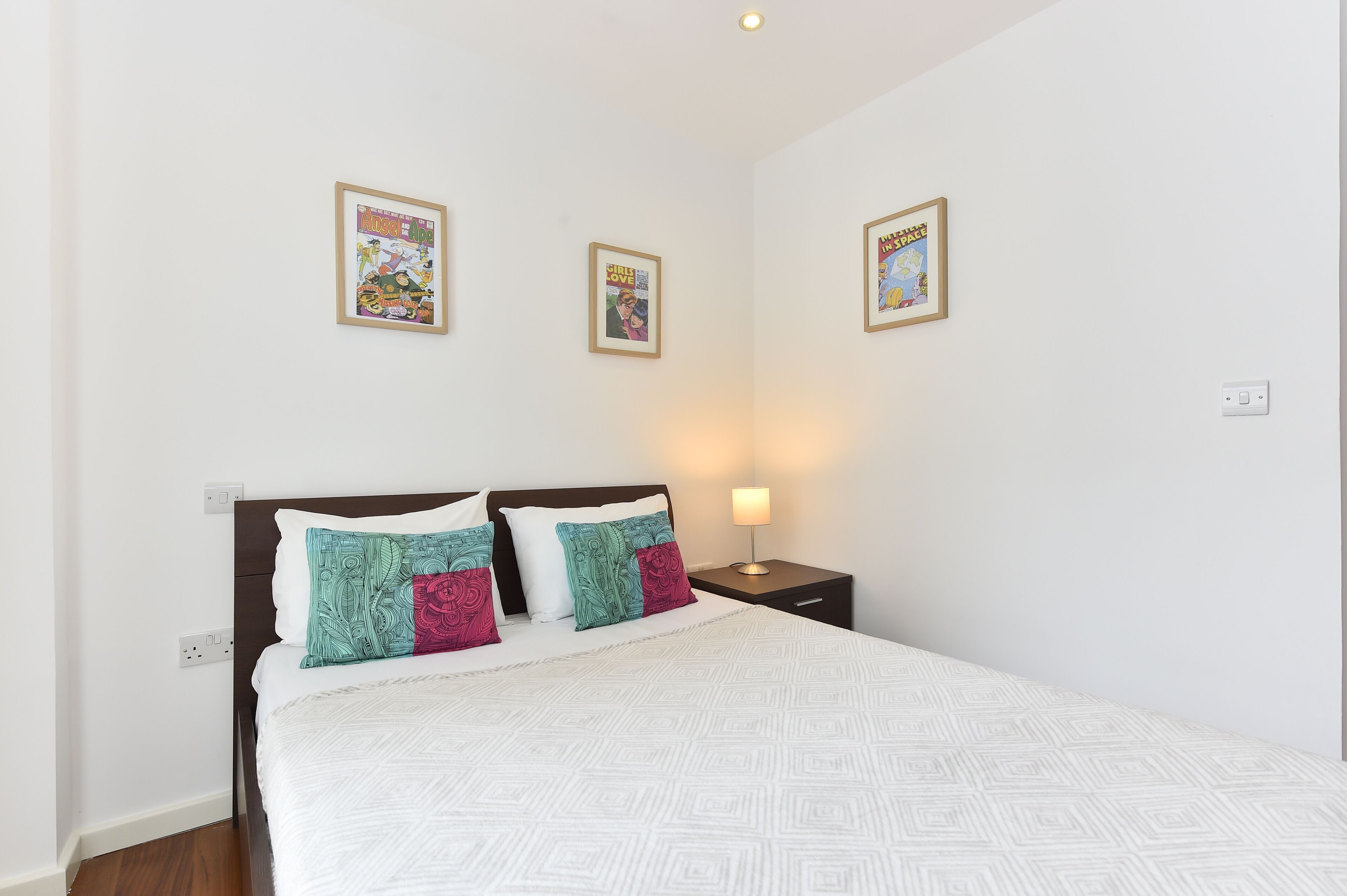 A neatly made bed with patterned bedding, a bedside table with a lamp, and three framed comic book posters on the wall.