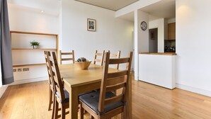A dining area with a wooden table and chairs, a small plant on a shelf, and a kitchen in the background. - Shoreditch Apartments (London)