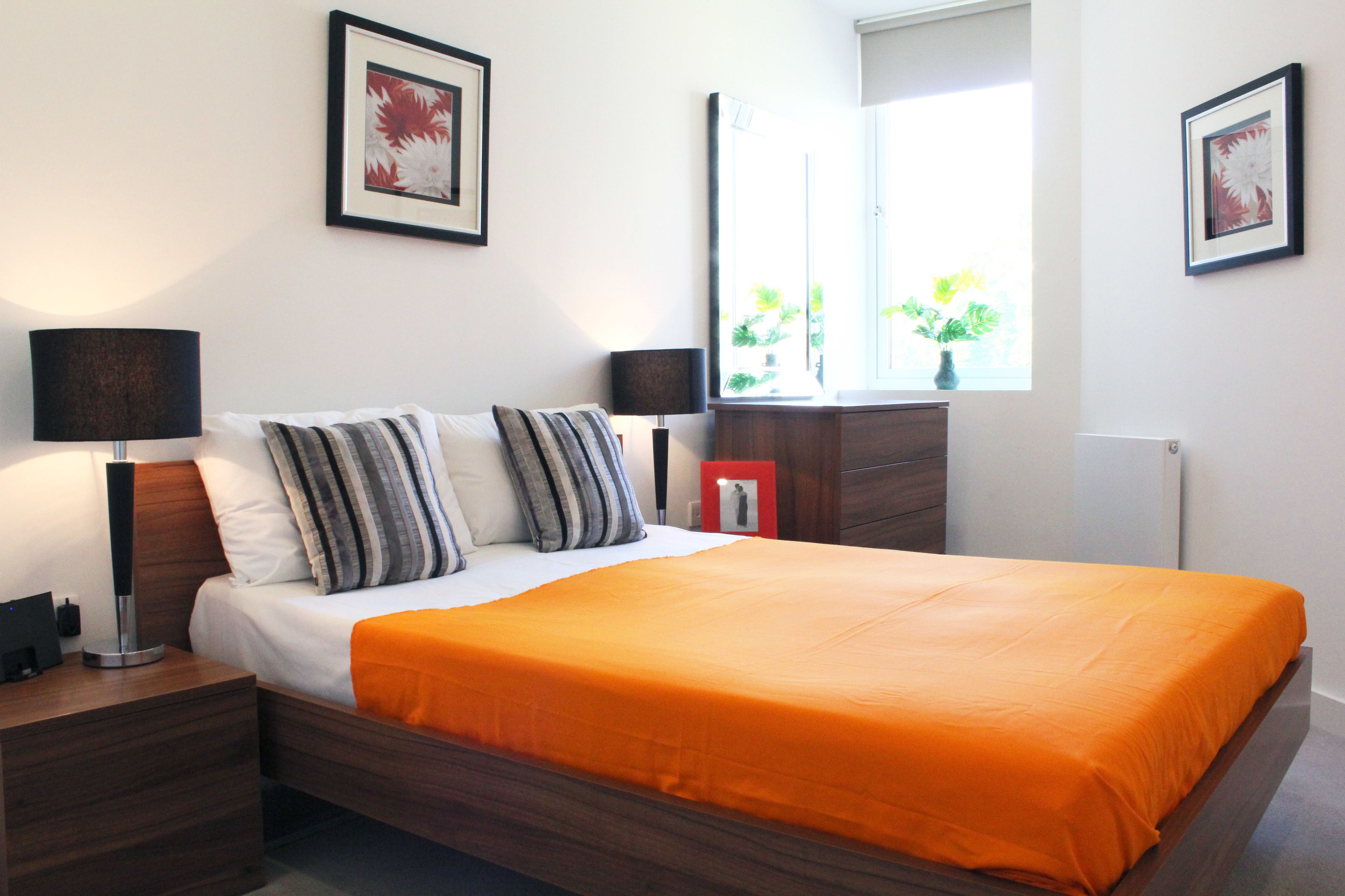 A neatly made bed with an orange bedspread, a bedside lamp, and a nightstand. There are two framed pictures on the wall above the bed.