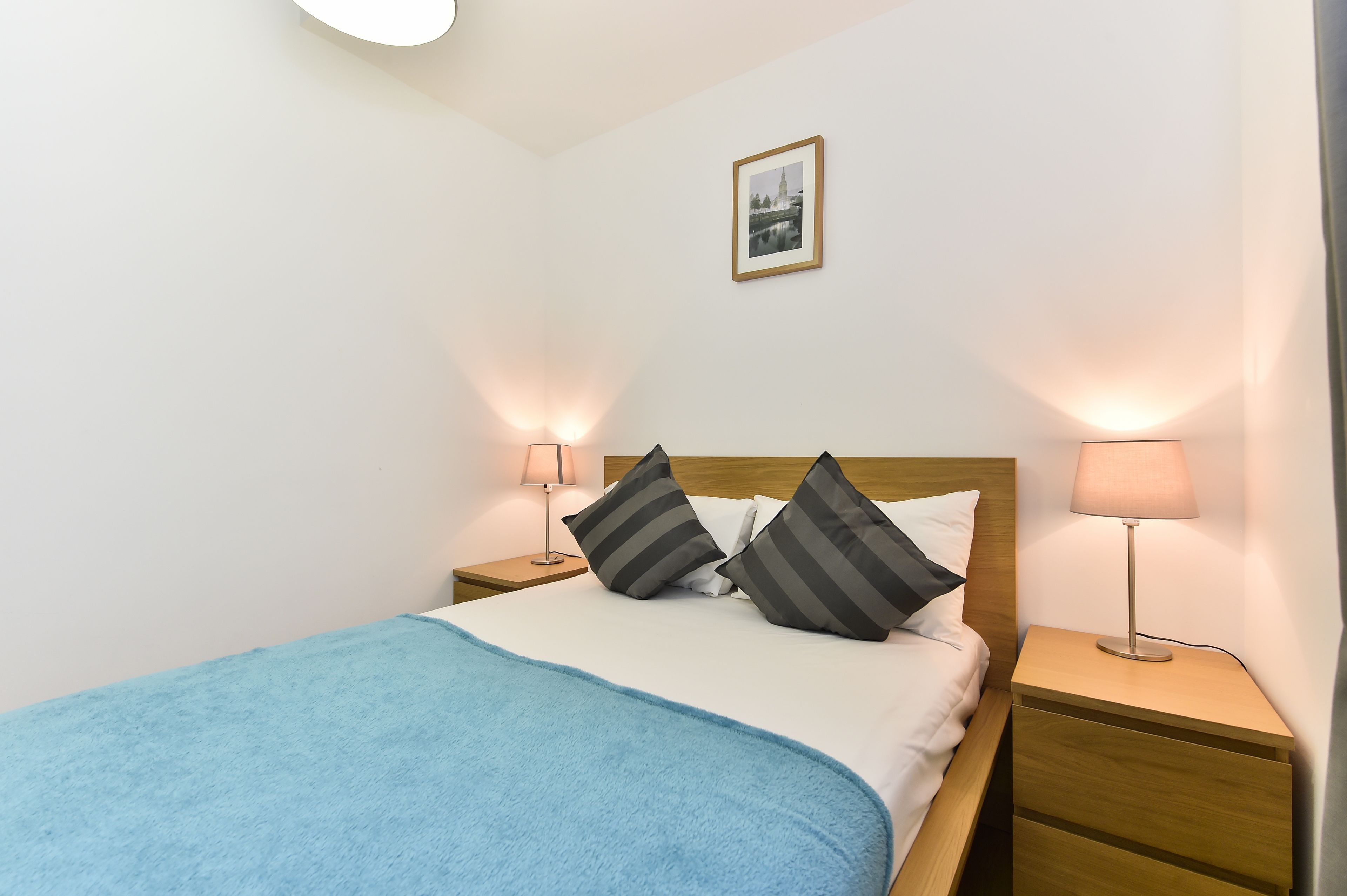 A neatly made bed with a blue blanket, two bedside lamps, and a framed picture on the wall.