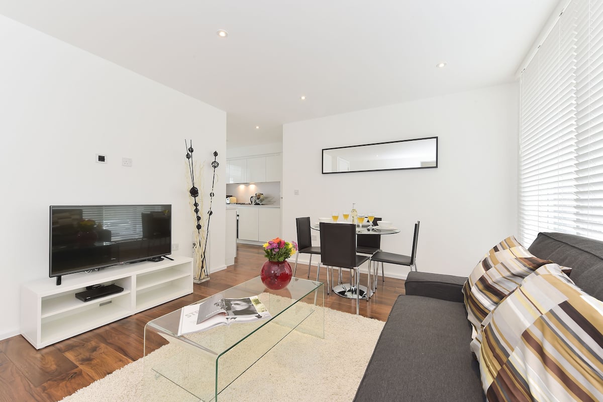A modern living room with a flat-screen TV, a glass coffee table, a sofa, and a dining area with a table and chairs.