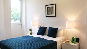 A neatly made bed with a blue blanket, two bedside lamps, a framed picture on the wall, and a small table with a vase and a book. - Shoreditch Apartments (London)
