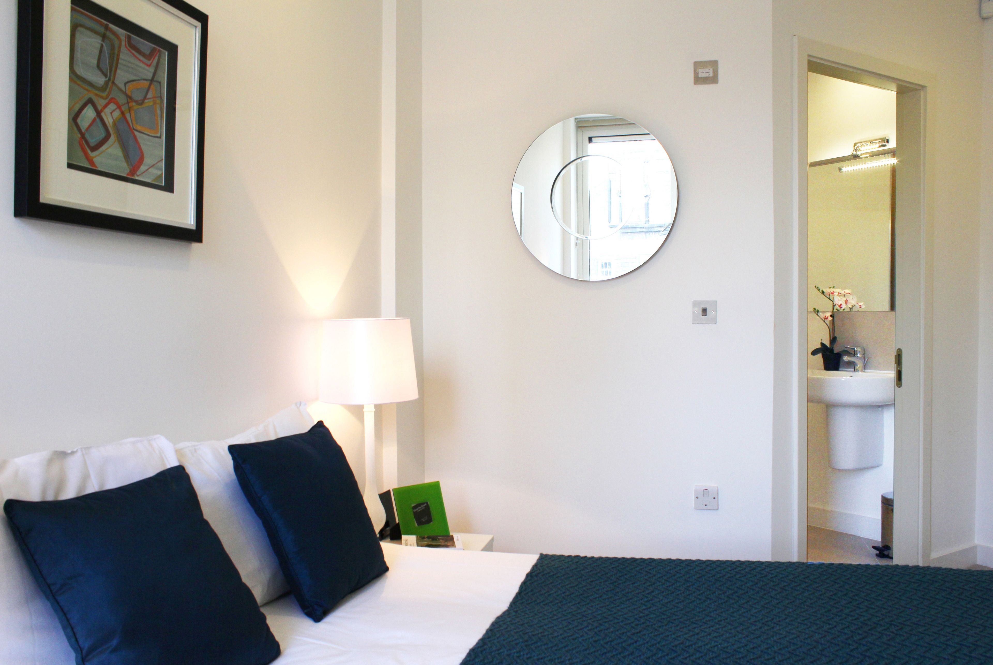 A hotel room with a bed, pillows, a bedside table with a lamp, a framed picture, a round mirror, and a view into the bathroom.