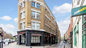 A corner building with a Subway sign, a cafe, and a street with cars and pedestrians. - Shoreditch Apartments (London)