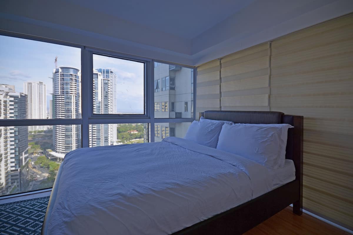 Een slaapkamer met een groot raam, een bed en uitzicht op hoge gebouwen.